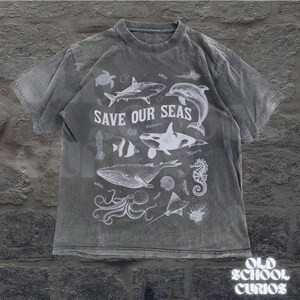 Puede incluir: Una camiseta gris con un aspecto desgastado, que presenta un gr&aacute;fico blanco de varias criaturas marinas, incluyendo una ballena, un tibur&oacute;n y un delf&iacute;n. El texto "SAVE OUR SEAS" se muestra prominentemente sobre las ilustraciones. La camiseta es de "OLD SCHOOL CURIOS".