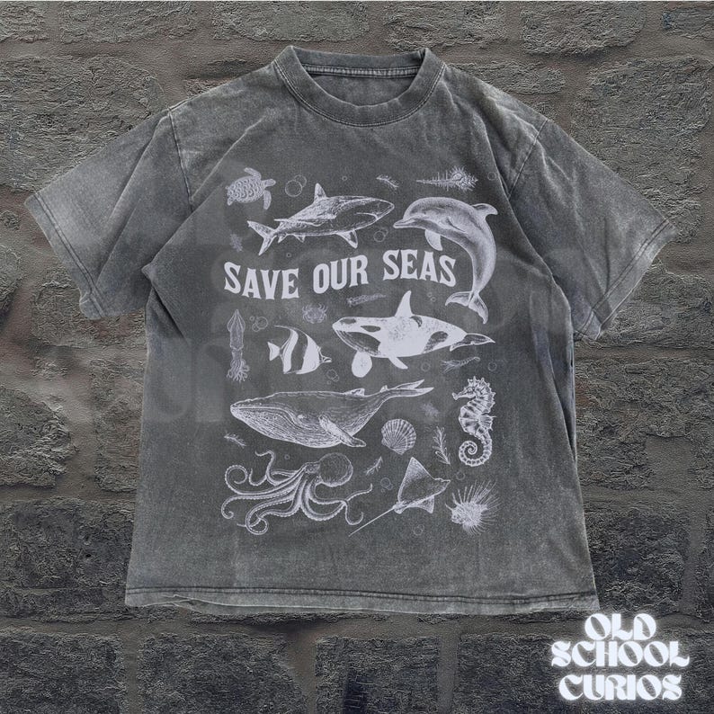 Puede incluir: Una camiseta gris con un aspecto desgastado, que presenta un gr&aacute;fico blanco de varias criaturas marinas, incluyendo una ballena, un tibur&oacute;n y un delf&iacute;n. El texto "SAVE OUR SEAS" se muestra prominentemente sobre las ilustraciones. La camiseta es de "OLD SCHOOL CURIOS".