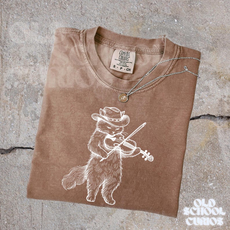 May include: Brown Comfort Colors t-shirt with a white illustration of a cat playing a violin and wearing a cowboy hat. A silver necklace with a small pendant rests on the shirt. The shirt is folded on a concrete surface.