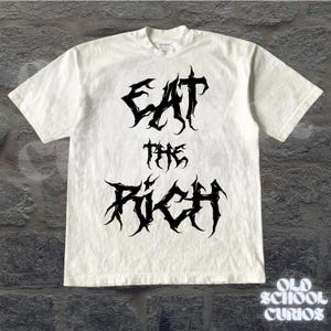 May include: White t-shirt with the text "EAT THE RICH" in a black, gothic font. The shirt has short sleeves and a crew neck. The brand name "Gildan" is visible on the collar.