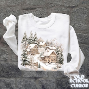 Op de afbeelding: Witte sweatshirt met een aquarel illustratie van met sneeuw bedekte huizen te midden van groenblijvende bomen. Het ontwerp toont een winterdorpssc&egrave;ne met een kronkelende weg. De tekst "OLD SCHOOL CURIOS" is gedrukt in een vintage lettertype.