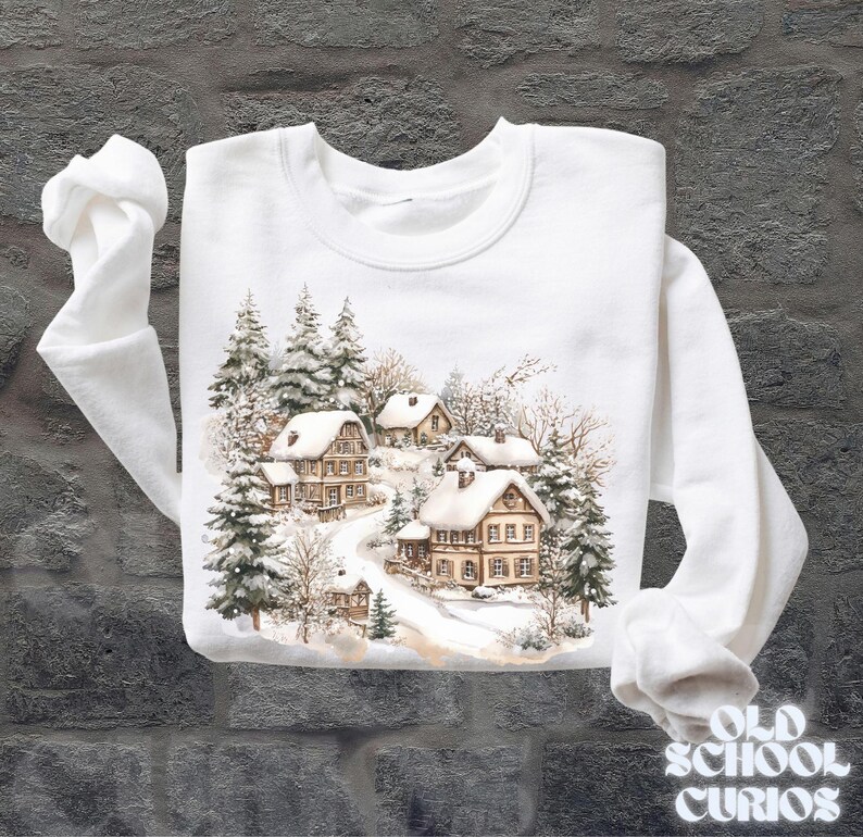 Op de afbeelding: Witte sweatshirt met een aquarel illustratie van met sneeuw bedekte huizen te midden van groenblijvende bomen. Het ontwerp toont een winterdorpssc&egrave;ne met een kronkelende weg. De tekst "OLD SCHOOL CURIOS" is gedrukt in een vintage lettertype.