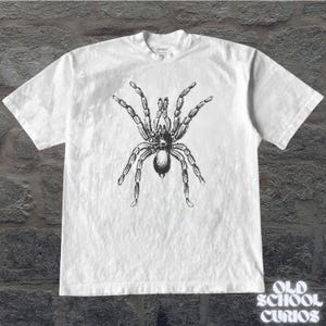 Peut inclure: T-shirt blanc avec une illustration d&eacute;taill&eacute;e en noir et gris d'une araign&eacute;e tarentule. Le t-shirt a un col rond et des manches courtes. Le texte "OLD SCHOOL CURIOS" est imprim&eacute; en blanc en bas &agrave; droite.