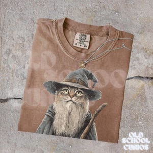 K&ouml;nnte beinhalten: Braunes Comfort Colors T-Shirt mit einer Katze als Zauberer mit langem wei&szlig;em Bart, blauem Hut und Robe, die einen Holzstab h&auml;lt. Eine silberne Halskette mit einem kleinen Anh&auml;nger liegt auf dem Shirt. Das Shirt hat den Text "Old School Curios".