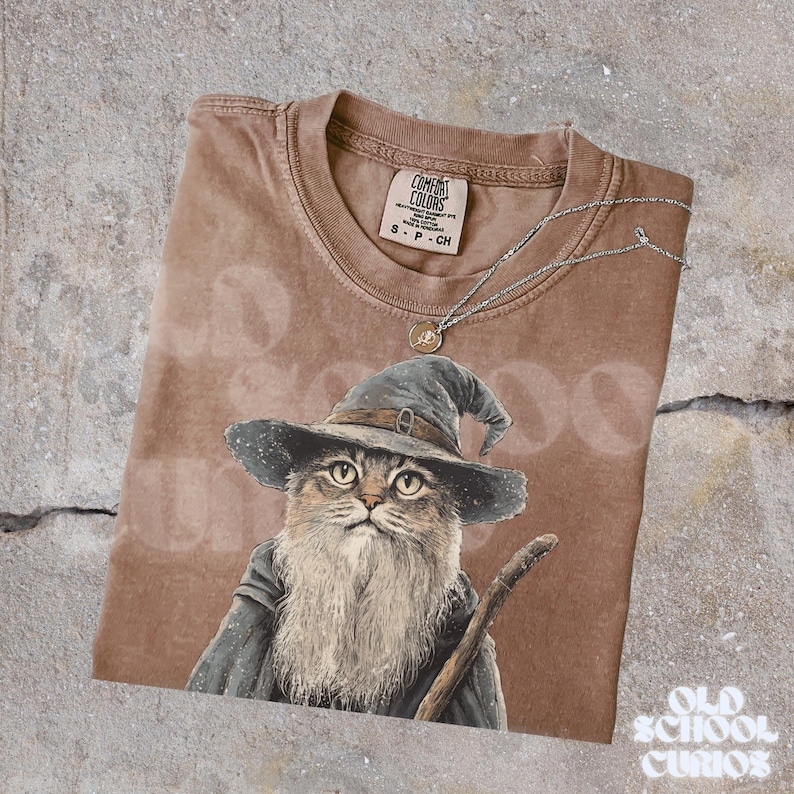 K&ouml;nnte beinhalten: Braunes Comfort Colors T-Shirt mit einer Katze als Zauberer mit langem wei&szlig;em Bart, blauem Hut und Robe, die einen Holzstab h&auml;lt. Eine silberne Halskette mit einem kleinen Anh&auml;nger liegt auf dem Shirt. Das Shirt hat den Text "Old School Curios".