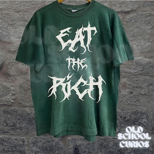 May include: Green t-shirt with the white text "EAT THE RICH" in a gothic font. The shirt has short sleeves and a crew neck. The words "OLD SCHOOL CURIOS" are in white text at the bottom right.