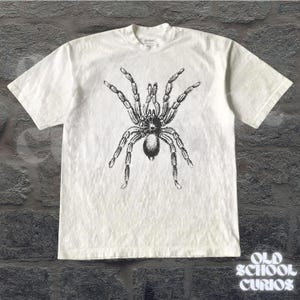 Peut inclure: T-shirt blanc avec une illustration d&eacute;taill&eacute;e en noir et gris d'une tarentule. Le t-shirt a un col rond et des manches courtes. Le texte "OLD SCHOOL CURIOS" est visible en bas &agrave; droite.