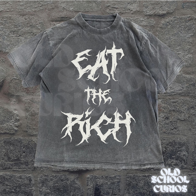 May include: A gray t-shirt with the text "EAT THE RICH" in a white, gothic font. The shirt has a vintage, washed appearance. The words are stacked vertically on the front of the shirt. The brand "OLD SCHOOL CURIOS" is printed at the bottom right.
