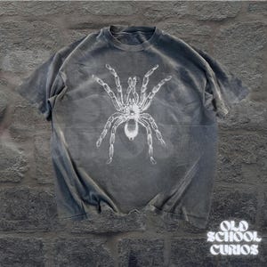 Peut inclure: T-shirt gris avec un motif blanc de tarentule. Le t-shirt a un aspect vintage et vieilli. Les mots "OLD SCHOOL CURIOS" sont imprim&eacute;s en blanc en bas &agrave; droite.