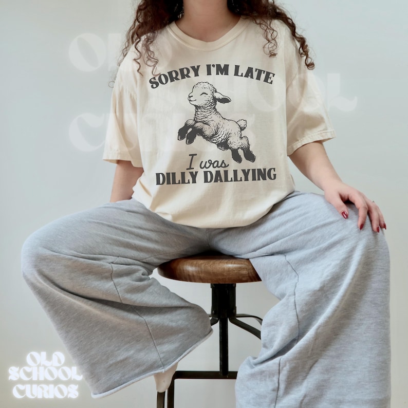 May include: Cream-colored t-shirt with the text "SORRY I'M LATE" above a jumping lamb illustration and the text "I was DILLY DALLYING". The shirt is paired with gray sweatpants. The overall theme is casual and humorous.
