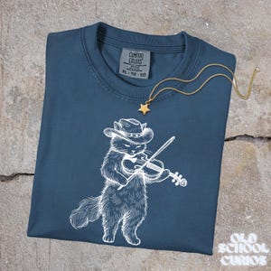 May include: A navy blue Comfort Colors t-shirt featuring a white illustration of a cat wearing a cowboy hat and playing a violin. A gold star pendant necklace rests on the shirt. The shirt is labeled XL-TG-EG.