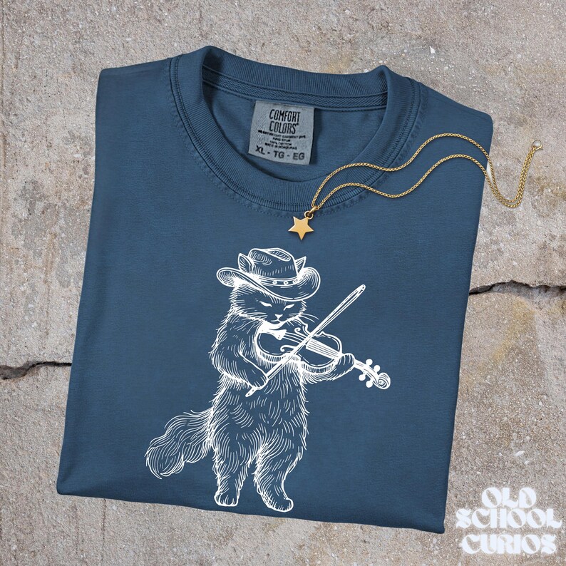 May include: A navy blue Comfort Colors t-shirt featuring a white illustration of a cat wearing a cowboy hat and playing a violin. A gold star pendant necklace rests on the shirt. The shirt is labeled XL-TG-EG.