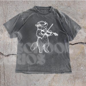 May include: A charcoal gray t-shirt featuring a white illustration of a cat wearing a cowboy hat and playing a violin. The shirt has a vintage, washed appearance. The word "SCHOOL" is faintly visible in the background.