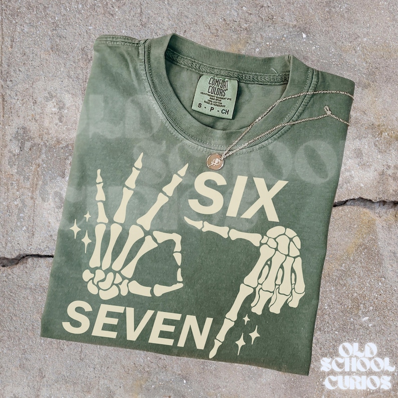 May include: An olive green t-shirt with a skeleton hand graphic displaying the numbers "SIX" and "SEVEN". A silver necklace with a small pendant rests on the shirt. The shirt is folded on a textured surface.