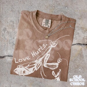 May include: Brown Comfort Colors t-shirt with a white praying mantis graphic and the words "Love Hurts." A silver necklace with a small pendant rests on the shirt. The shirt has a small tag.