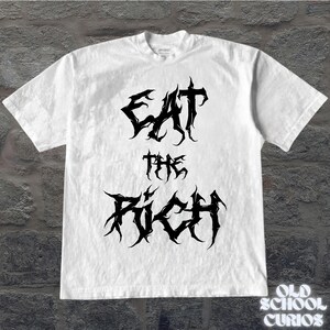 May include: White t-shirt with the text "EAT THE RICH" in a spiky, gothic font. The shirt is plain and has a crew neck. The image is set against a textured, gray background.
