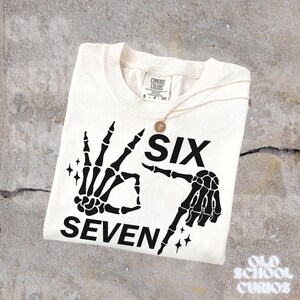 May include: Cream-colored t-shirt with a black skeleton hand graphic forming the numbers six and seven. The shirt includes the text "SIX SEVEN" and is paired with a gold necklace. The shirt is from Comfort Colors.