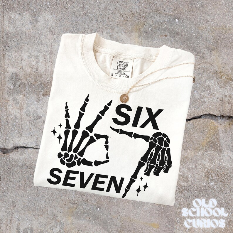 May include: Cream-colored t-shirt with a black skeleton hand graphic forming the numbers six and seven. The shirt includes the text "SIX SEVEN" and is paired with a gold necklace. The shirt is from Comfort Colors.