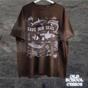 Puede incluir: Camiseta marr&oacute;n con un gr&aacute;fico blanco de varias criaturas marinas, incluyendo una ballena, un tibur&oacute;n y un delf&iacute;n. El texto "SAVE OUR SEAS" est&aacute; impreso encima de las ilustraciones. La camiseta est&aacute; colgada en una percha blanca.