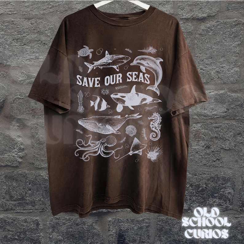 Puede incluir: Camiseta marr&oacute;n con un gr&aacute;fico blanco de varias criaturas marinas, incluyendo una ballena, un tibur&oacute;n y un delf&iacute;n. El texto "SAVE OUR SEAS" est&aacute; impreso encima de las ilustraciones. La camiseta est&aacute; colgada en una percha blanca.