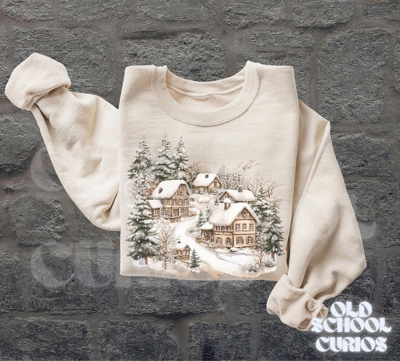 Op de afbeelding: Een cr&egrave;mekleurige sweatshirt met een winterdorpssc&egrave;ne, met besneeuwde huizen en groenblijvende bomen. De afbeelding toont een besneeuwde weg die door het dorp slingert. De tekst "OLD SCHOOL CURIOS" is rechtsonder zichtbaar.