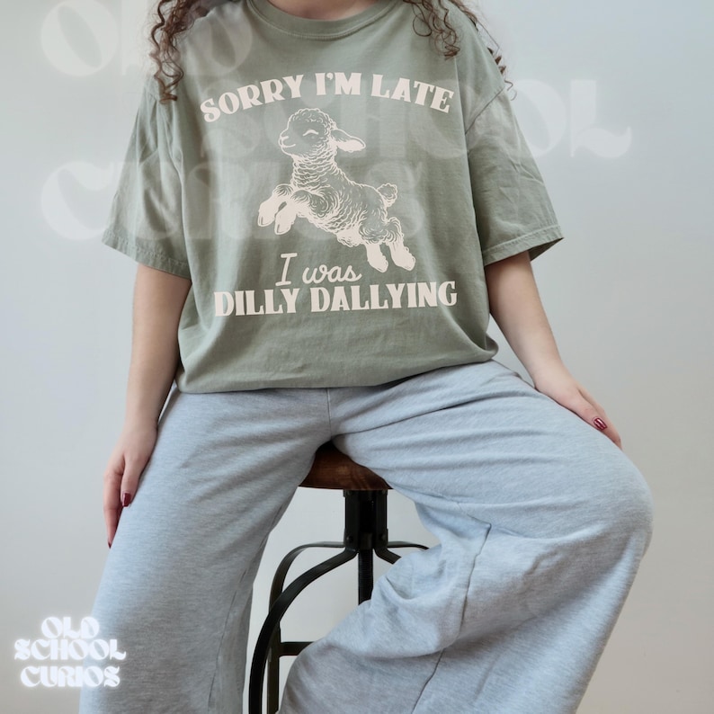 May include: Sage green t-shirt with the text "SORRY I'M LATE" above a leaping lamb illustration and the text "I WAS DILLY DALLYING". The shirt is paired with gray sweatpants. The model is seated on a stool.