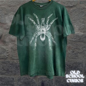 Peut inclure: T-shirt vert avec un motif d'araign&eacute;e blanche. Le t-shirt a un aspect vintage et d&eacute;lav&eacute;. Le texte "OLD SCHOOL CURIOS" est imprim&eacute; en blanc dans le coin inf&eacute;rieur droit.