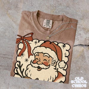 May include: A brown Comfort Colors t-shirt featuring a vintage Santa Claus graphic with a red hat and beard. The design includes a red bow and snowflakes. A silver necklace with a pendant rests on the shirt. The words "Old School Curios" are at the bottom right.