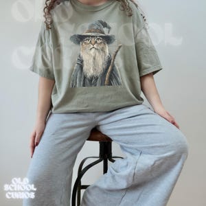 K&ouml;nnte beinhalten: Salbeigr&uuml;nes T-Shirt mit einer detaillierten Illustration einer Katze, die als Zauberer gekleidet ist, komplett mit Hut, Robe und Stab. Die Katze hat einen langen wei&szlig;en Bart. Das Shirt hat eine lockere Passform.