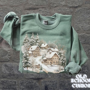 Op de afbeelding: Een saliegroene sweatshirt met een winterdorpssc&egrave;ne, met besneeuwde huizen en bomen. De afbeelding toont een kronkelende weg door het dorp. De woorden "OLD SCHOOL CURIOS" staan rechtsonder in het wit gedrukt.