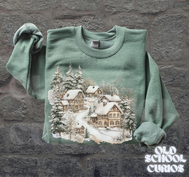 Op de afbeelding: Een saliegroene sweatshirt met een winterdorpssc&egrave;ne, met besneeuwde huizen en bomen. De afbeelding toont een kronkelende weg door het dorp. De woorden "OLD SCHOOL CURIOS" staan rechtsonder in het wit gedrukt.