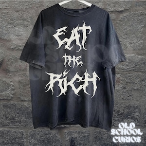 May include: Black t-shirt with the text "EAT THE RICH" in a white, gothic font. The shirt is hanging on a white hanger against a textured wall. The words "OLD SCHOOL CURIOS" are at the bottom.