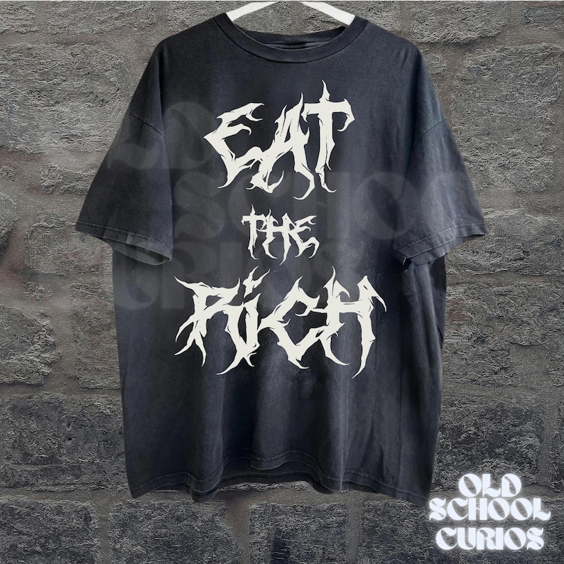 May include: Black t-shirt with the text "EAT THE RICH" in a white, gothic font. The shirt is hanging on a white hanger against a textured wall. The words "OLD SCHOOL CURIOS" are at the bottom.