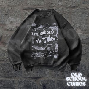 Puede incluir: Sudadera gris oscuro con un gr&aacute;fico blanco de varias criaturas marinas, incluyendo una ballena, un tibur&oacute;n y un delf&iacute;n. El texto "SAVE OUR SEAS" se muestra de forma destacada. La sudadera tambi&eacute;n presenta el texto "OLD SCHOOL CURIOS".