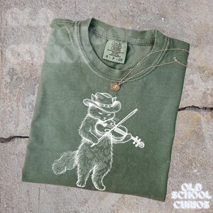 May include: Olive green t-shirt featuring a white illustration of a cat wearing a cowboy hat and playing a violin. A gold-colored necklace with a pendant rests on the shirt. The shirt has a Comfort Colors tag.