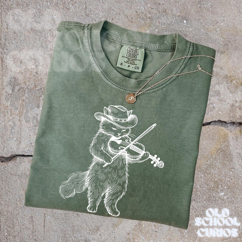 May include: Olive green t-shirt featuring a white illustration of a cat wearing a cowboy hat and playing a violin. A gold-colored necklace with a pendant rests on the shirt. The shirt has a Comfort Colors tag.