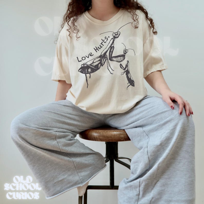 May include: A cream-coloured t-shirt featuring a black graphic of praying mantises and the text "Love Hurts." The shirt is worn with grey sweatpants. The image is from Old School Curios.