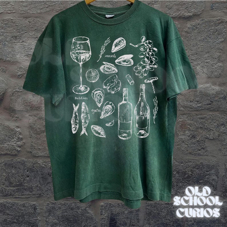 May include: Green t-shirt with a white illustration of wine, sardines, oysters, mussels, and wine bottles. The text "Old School Curios" is in the bottom right corner. The shirt is made of a soft material.