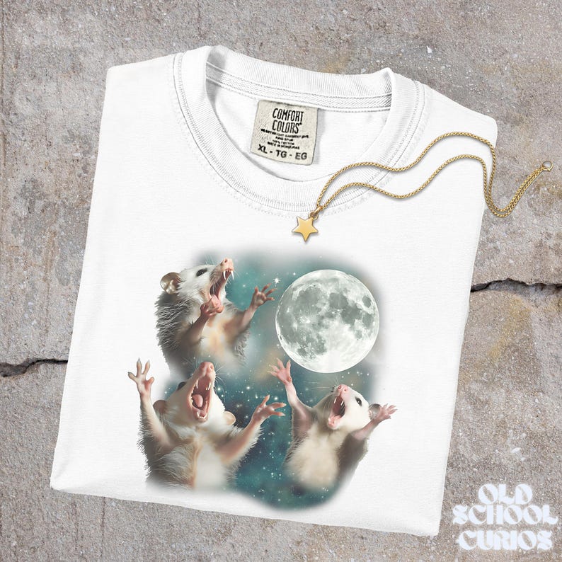 K&ouml;nnte beinhalten: Wei&szlig;es Comfort Colors T-Shirt mit einer Grafik von drei Opossums, die einen Vollmond vor einem Sternenhimmel anheulen. Eine goldfarbene Halskette mit einem Sternanh&auml;nger liegt auf dem Shirt. Das Etikett des Shirts lautet XL-TG-EG. Der Text "Old School Curios" befindet sich unten rechts.