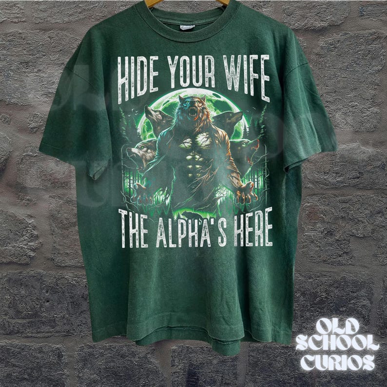 May include: Green t-shirt with the text "HIDE YOUR WIFE THE ALPHA'S HERE" in white. The graphic features a werewolf with three wolf heads in front of a full moon. The shirt has a vintage, distressed look.