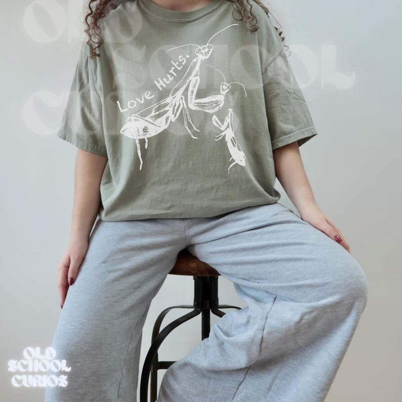 May include: Sage green t-shirt featuring a white graphic of two praying mantises and the text "Love Hurts." The shirt is worn with grey sweatpants. The style is casual and relaxed.