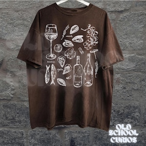 May include: Brown t-shirt with a white illustration of seafood and wine elements, including sardines, oysters, mussels, wine bottles, and a wine glass. The shirt has the text "Old School Curios" at the bottom.