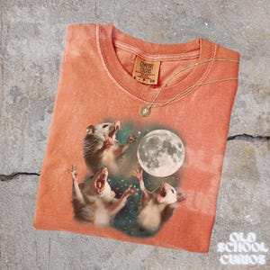 K&ouml;nnte beinhalten: Korallfarbenes T-Shirt mit einer Grafik von drei Opossums, die den Vollmond anheulen. Das Shirt hat einen Rundhalsausschnitt und kurze &Auml;rmel. Eine goldene Halskette mit Anh&auml;nger liegt auf dem Shirt. Das Markenetikett lautet "Comfort Colors."