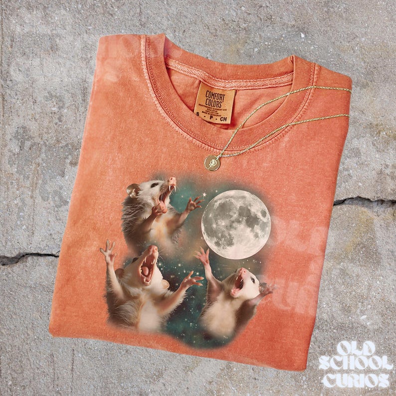 K&ouml;nnte beinhalten: Korallfarbenes T-Shirt mit einer Grafik von drei Opossums, die den Vollmond anheulen. Das Shirt hat einen Rundhalsausschnitt und kurze &Auml;rmel. Eine goldene Halskette mit Anh&auml;nger liegt auf dem Shirt. Das Markenetikett lautet "Comfort Colors."