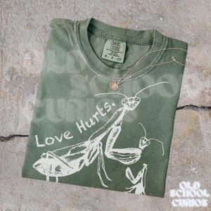 May include: An olive green t-shirt featuring a white praying mantis design and the words "Love Hurts." A silver necklace with a small pendant is draped across the shirt. The shirt is folded on a concrete surface.