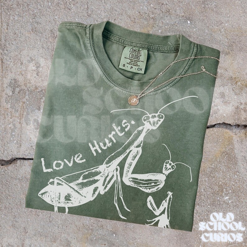May include: An olive green t-shirt featuring a white praying mantis design and the words "Love Hurts." A silver necklace with a small pendant is draped across the shirt. The shirt is folded on a concrete surface.