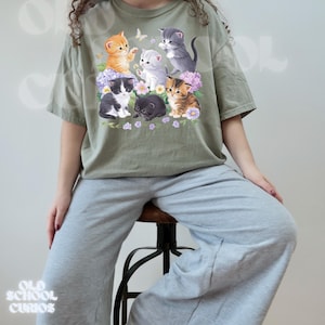以下が含まれることがあります： 花と蝶の中にいる7匹の子猫のグラフィックが特徴のオリーブグリーンのTシャツ。子猫はオレンジ、グレー、黒、茶色など様々な色です。シャツはグレーのスウェットパンツと組み合わせています。「OLD SCHOOL CURIOS」の文字が下にあります。
