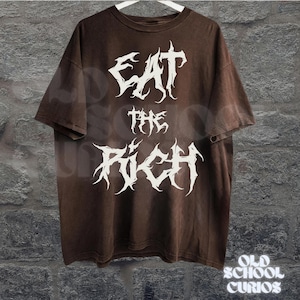 May include: Brown t-shirt with the text "EAT THE RICH" in a white, gothic font. The shirt has a vintage, faded look and is hanging on a white hanger against a textured wall. The words "OLD SCHOOL CURIOS" are at the bottom.