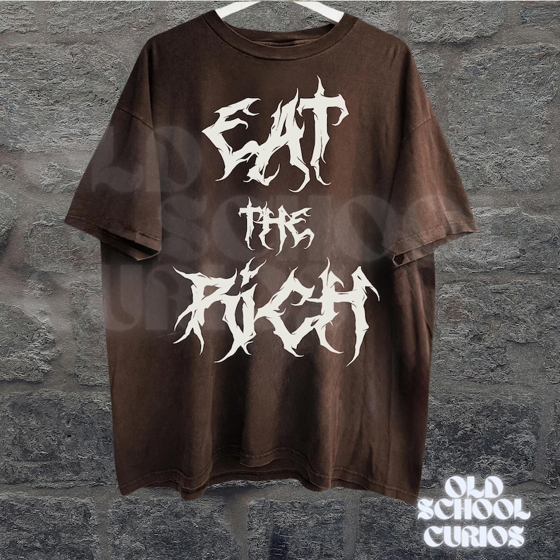 May include: Brown t-shirt with the text "EAT THE RICH" in a white, gothic font. The shirt has a vintage, faded look and is hanging on a white hanger against a textured wall. The words "OLD SCHOOL CURIOS" are at the bottom.
