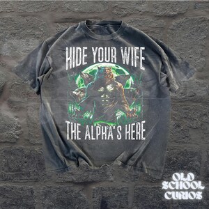 May include: Dark gray t-shirt with a werewolf graphic. The shirt features the text "HIDE YOUR WIFE THE ALPHA'S HERE" in white. The graphic includes a werewolf, wolves, and a green moon.